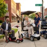 Two seniors using different mobility scooters on a sunny American sidewalk with accessories and a removable battery charging at a bench